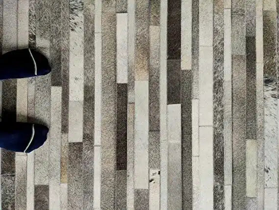 A pair of feet in dark socks with a light stripe stand on the RAMSHA HOME Grey Color Kitchen Decor Leather Rug - LR 004, featuring gray, beige, and brown rectangular tiles arranged in vertical stripes.