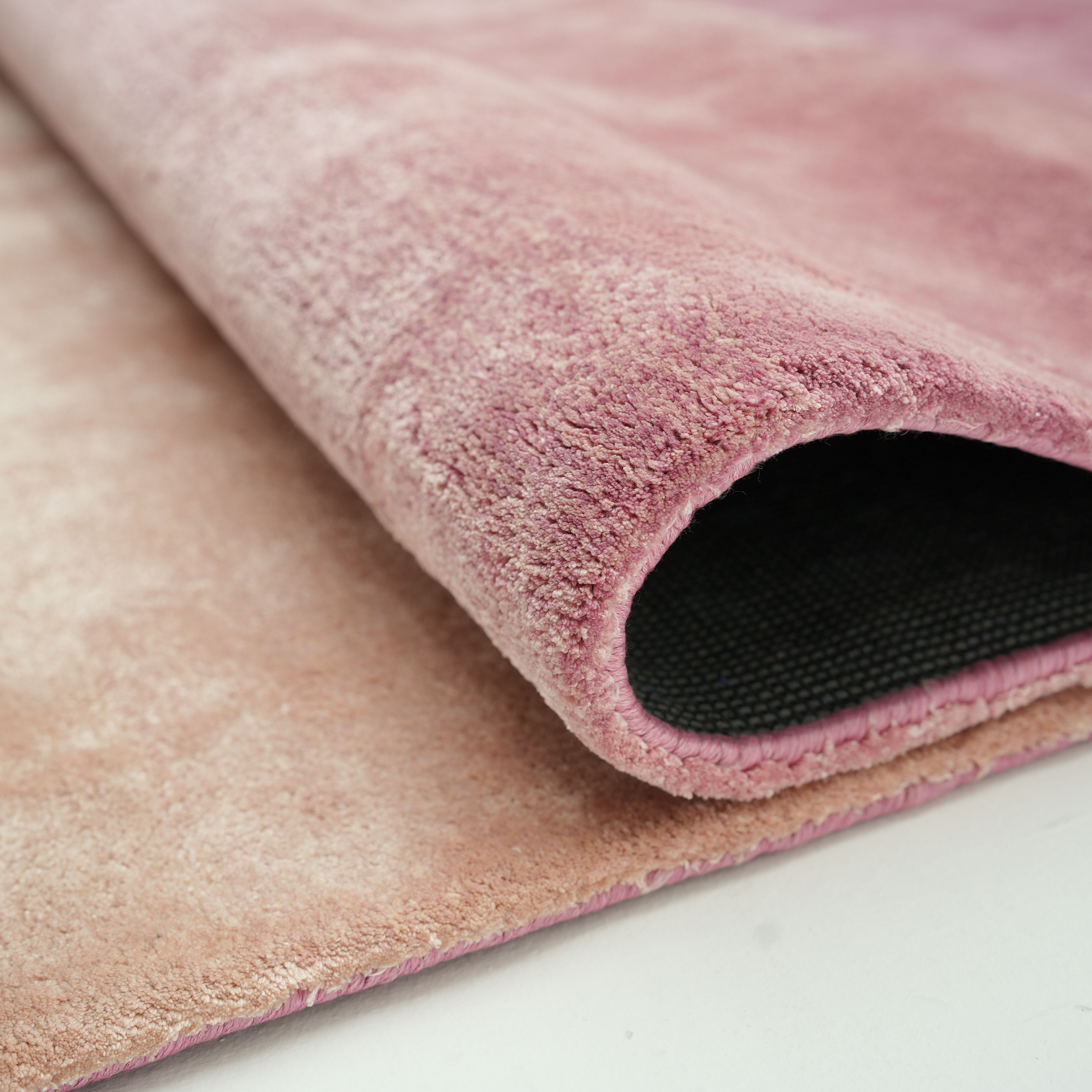 A close-up of the Rose Ombre Handmade Area Rug HTV 27 (300x400 cm) by RAMSHA HOME shows its soft pink surface and black textured backing, partially rolled on a light surface.