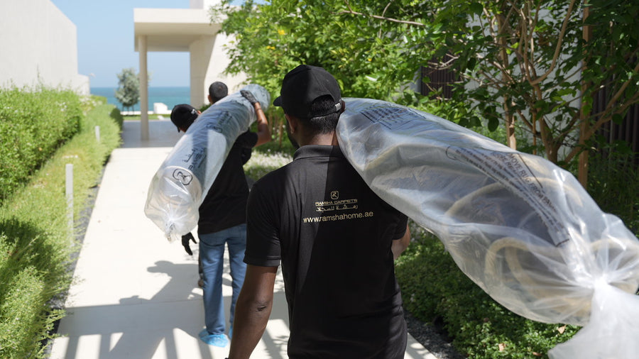 Two movers in black uniforms carry a large, plastic-wrapped RAMSHA HOME Handmade Rose Shape Rug HTW 106 on their shoulders along a garden path toward a modern building with a sea view in the background.