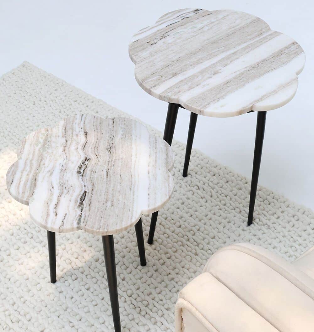 The CloudForm Nesting Marble Tables by RAMSHA HOME, featuring flower-shaped white marble tops and black legs, sit on a textured cream rug beside a beige upholstered chair.