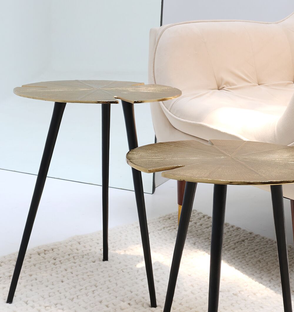 The Lily Luxe Aluminum Accent Tables – Set of 2 Nesting Tables by RAMSHA HOME, with black and gold leaf-shaped tops, stand on a white textured rug beside a cream upholstered armchair in a modern minimalist room.