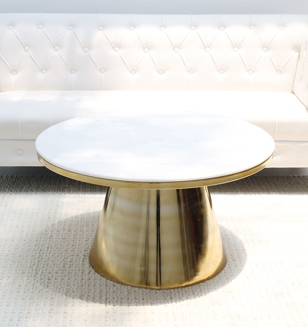The RAMSHA HOME Aurelia Luxe Marble & Iron Accent Table AD25-27, featuring a round white marble top and gleaming gold cone-shaped base, is placed on a textured light rug in front of a white tufted sofa.