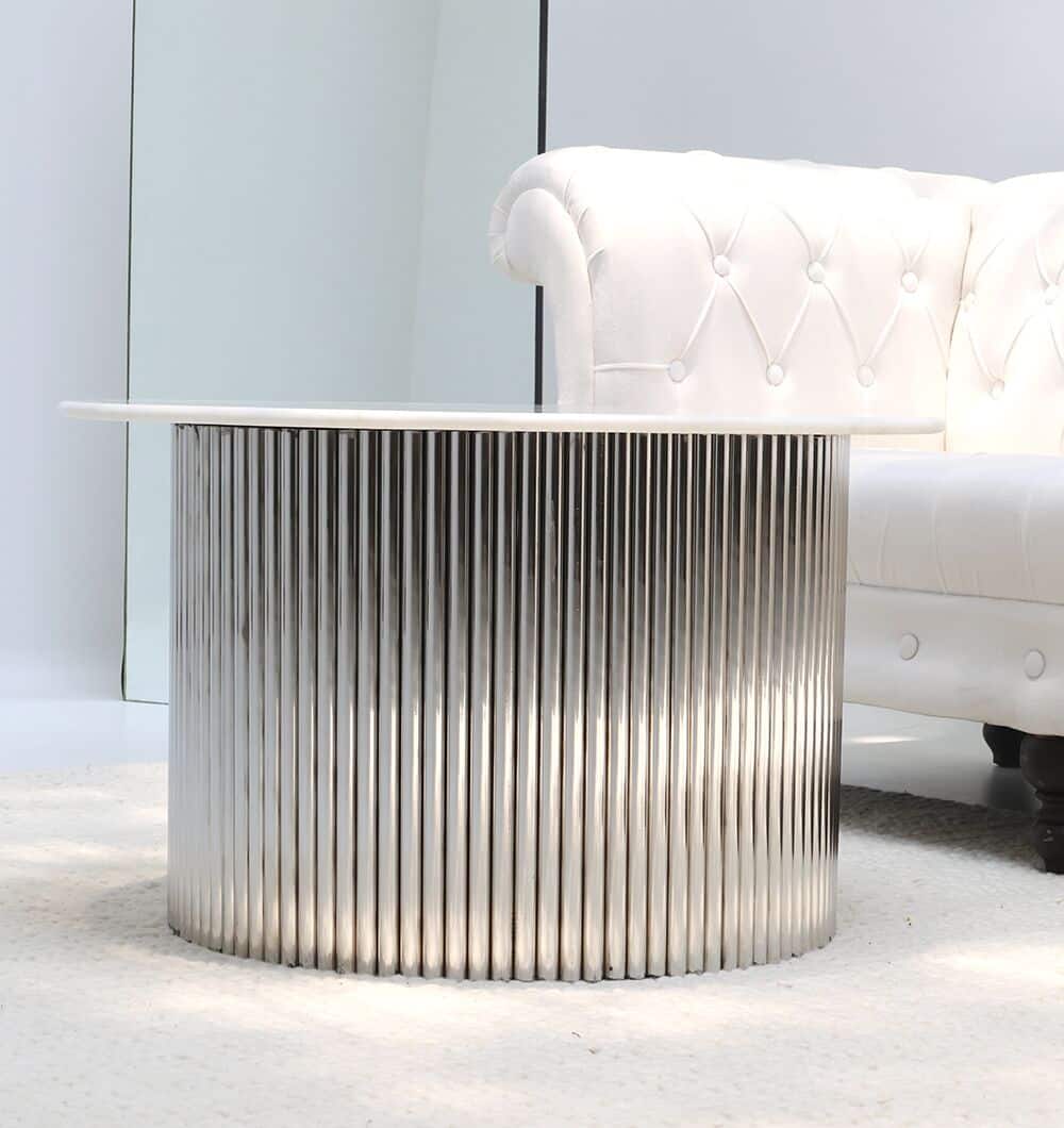 The Luna Polished Marble & Steel Coffee Table by RAMSHA HOME, featuring a ribbed stainless-steel base and smooth white top, sits next to a white tufted sofa on a light-colored carpet.