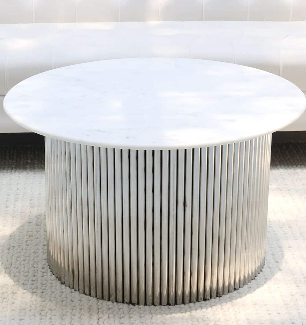 The RAMSHA HOME Luna Polished Marble & Steel Coffee Table features a round white marble top and cylindrical stainless-steel base with vertical slats, shown on a textured white rug in front of a white sofa.