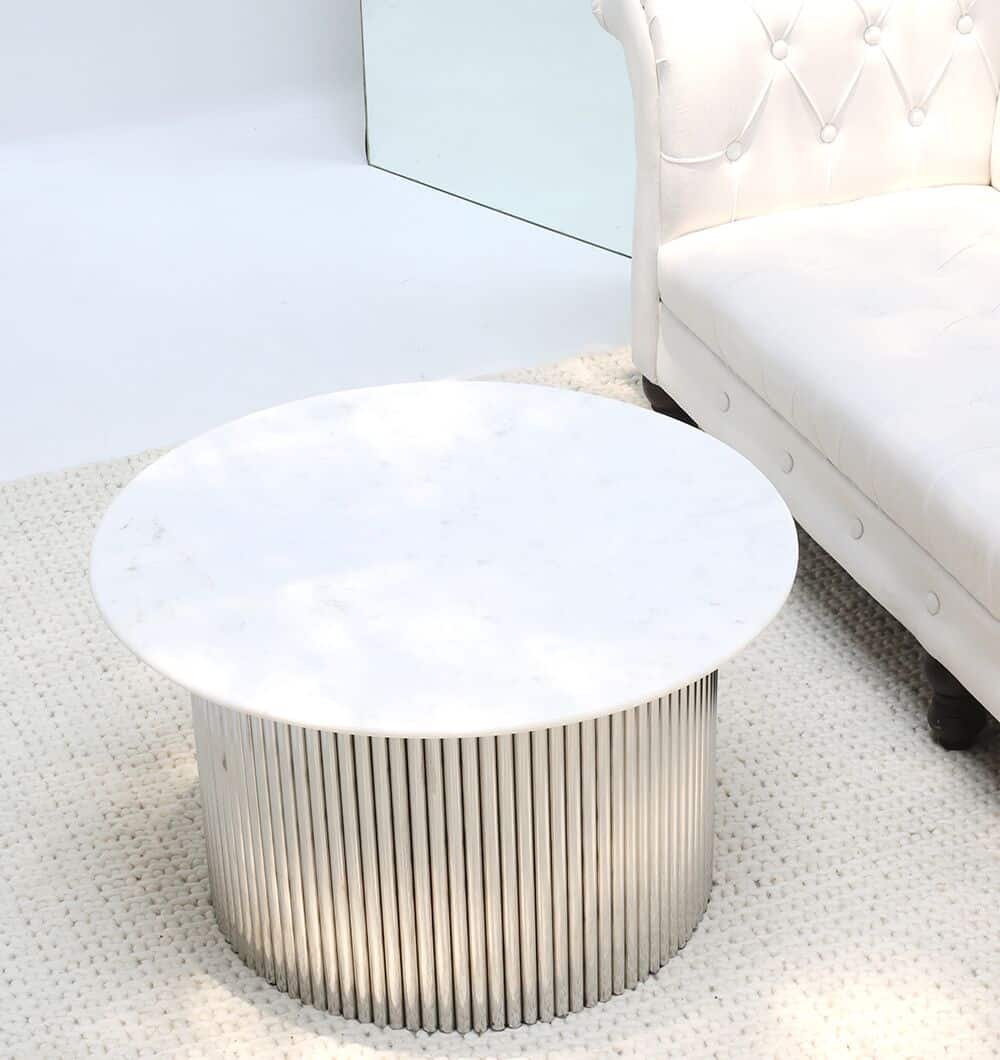 The Luna Polished Marble & Steel Coffee Table by RAMSHA HOME, featuring a white marble top and ribbed stainless-steel base, sits on a textured white rug beside a tufted white sofa in a bright, modern space.