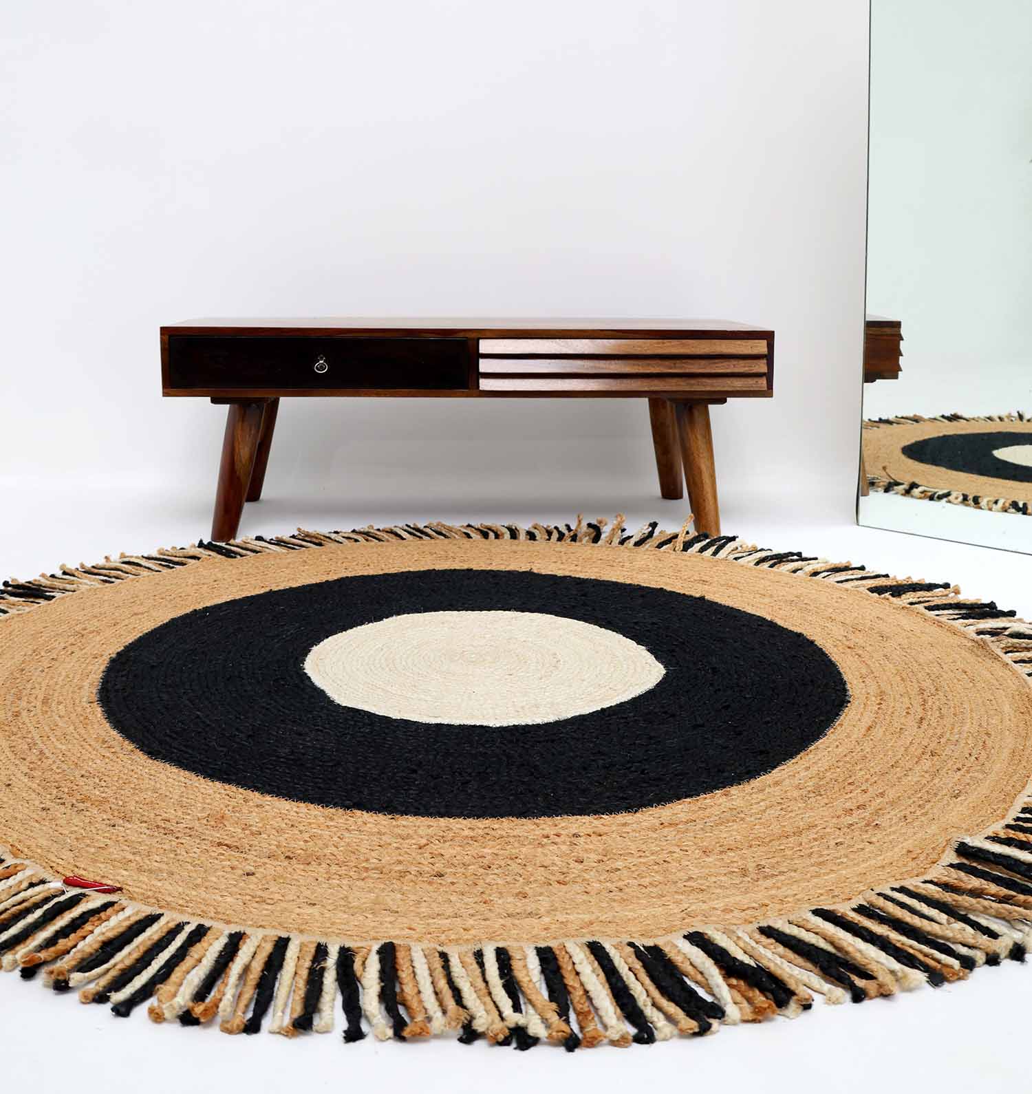 The RAMSHA HOME Amazing Handmade Braided Round Jute Rug - BR 001, featuring concentric beige, black, and cream circles with fringe edges, sits on a white floor before a wooden bench and is reflected in a mirror on the right.