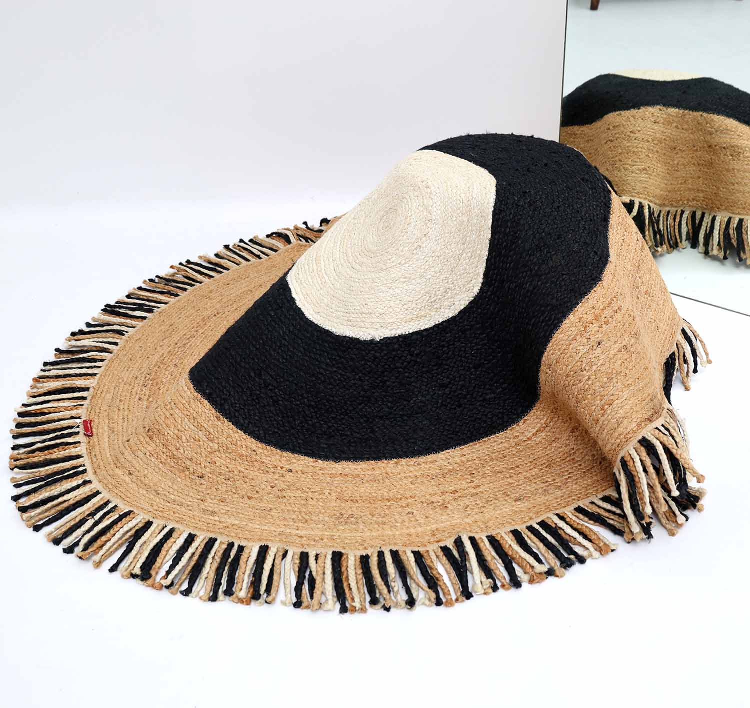 A large, woven straw hat with a wide brim and fringed edge sits beside the RAMSHA HOME Amazing Handmade Braided Round Jute Rug - BR 001, both resting on a white surface with part of the hat reflected in a mirror.