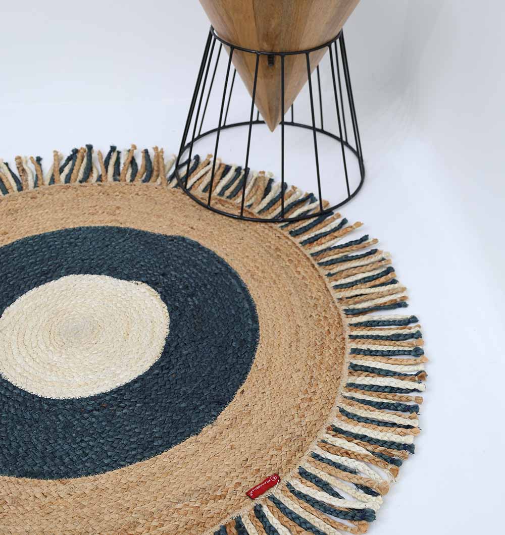 The RAMSHA HOME Amazing Handmade Braided Round Jute Rug - BR 001, with concentric beige, dark blue, and off-white circles and fringed edges, lies beside a wooden stool with black metal legs on a white floor.
