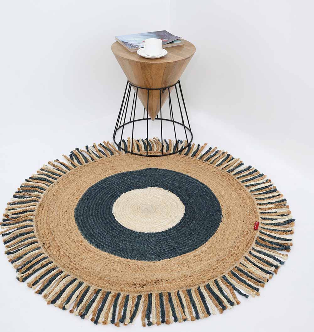 The RAMSHA HOME Amazing Handmade Braided Round Jute Rug - BR 001 with a fringed edge and cream, dark blue, and tan concentric circles sits on the floor beside a modern wooden side table holding books and a white cup.