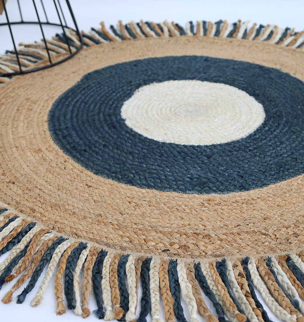 The RAMSHA HOME Amazing Handmade Braided Round Jute Rug - BR 001 features a central white circle, dark blue and tan rings, colorful fringe, and a glimpse of a black metal object in the corner.