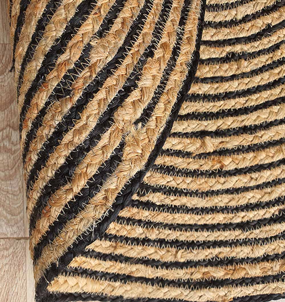 A close-up of the Natural & Black Color Braided Rug (Eco Friendly Jute Carpet BR-004) featuring alternating black and tan stripes, slightly curled at the edge, placed on a light wooden floor.