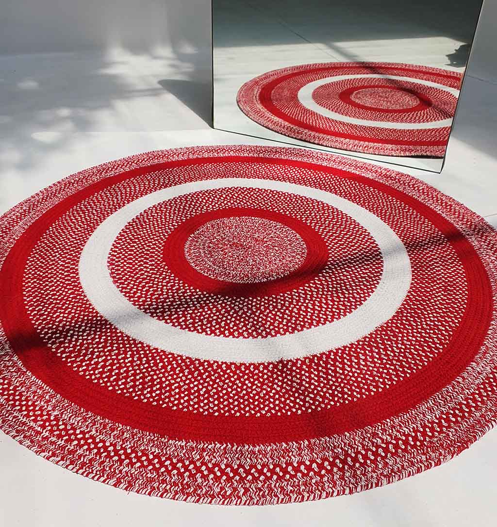The RAMSHA HOME Red & White Color Round Braided Rug - BR 005, featuring patterned concentric circles, is laid on a white floor and reflected in a large mirror behind it as leaf shadows fall across the scene.