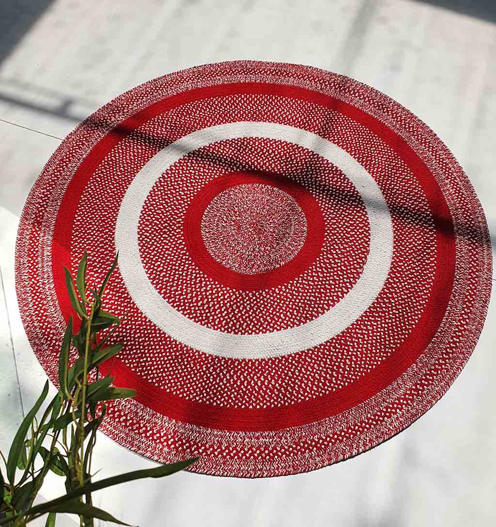 The RAMSHA HOME Red & White Color Round Braided Rug - BR 005, featuring concentric red and white circles and a textured pattern, is displayed on a light floor with green leafy plants partially visible in the lower left corner.