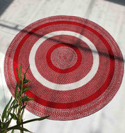 The RAMSHA HOME Red & White Color Round Braided Rug - BR 005, featuring concentric red and white circles and a textured pattern, is displayed on a light floor with green leafy plants partially visible in the lower left corner.