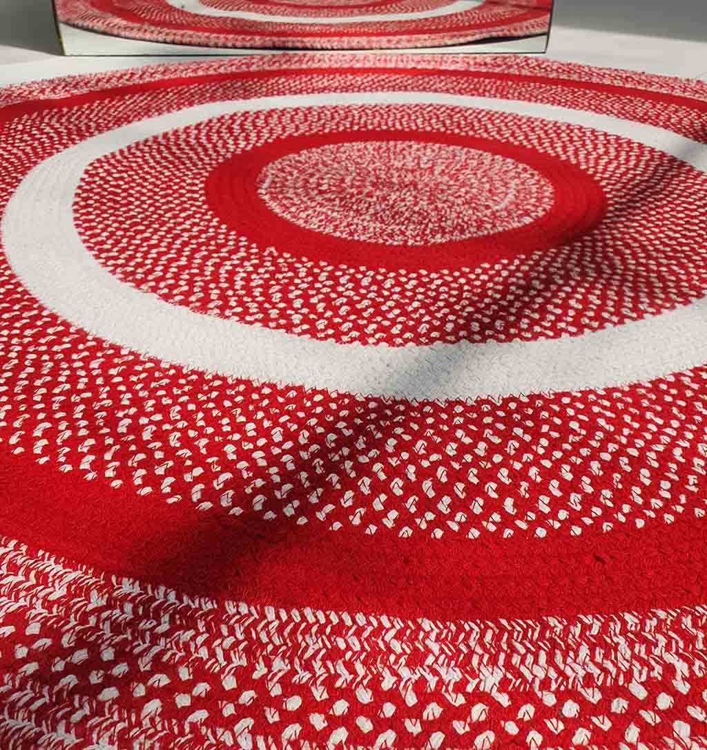 A close-up of the RAMSHA HOME Red & White Color Round Braided Rug - BR 005, featuring alternating concentric rings and a textured woven pattern, beautifully reflected in a nearby mirror.
