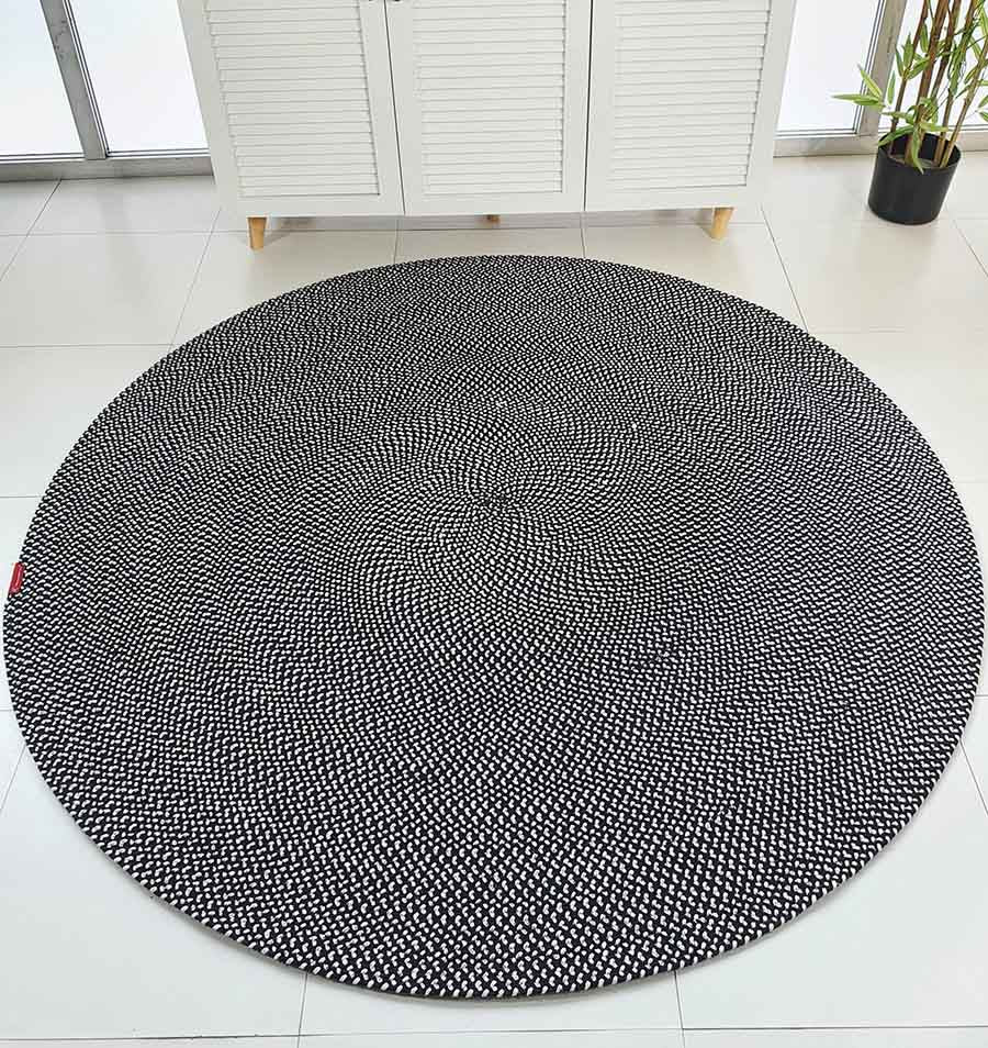 The RAMSHA HOME Black & White Color Round Braided Rug - BR 007, featuring a hypnotic optical illusion design, is displayed on a white tiled floor in a modern room with a white cabinet and a potted plant.