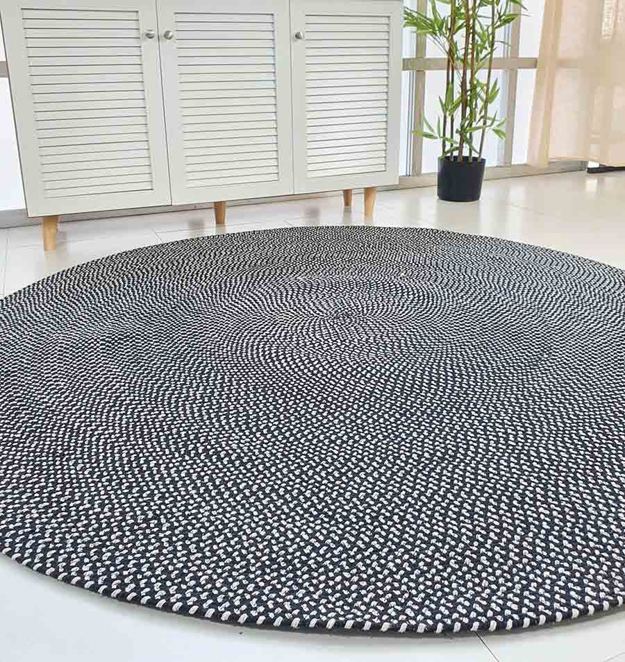 The RAMSHA HOME Black & White Color Round Braided Rug - BR 007 is displayed on a white tile floor in a bright room featuring a white cabinet, potted bamboo plant, and large windows.