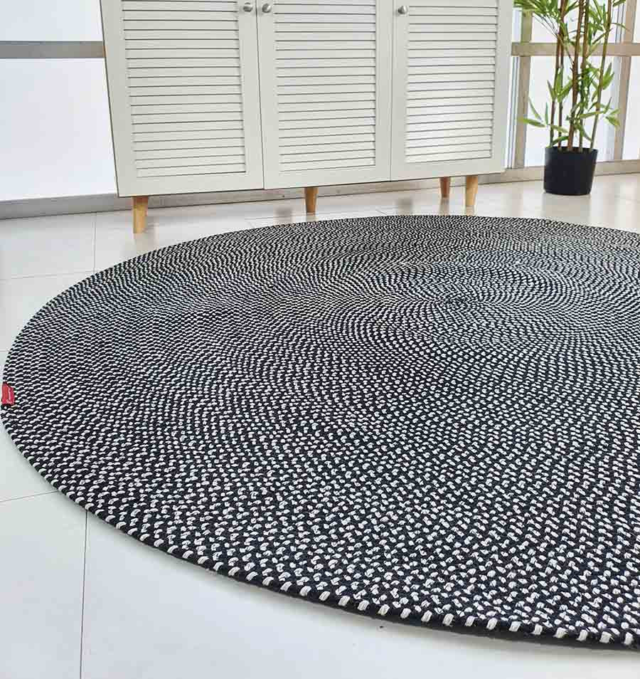 The RAMSHA HOME Black & White Color Round Braided Rug - BR 007, featuring a dotted pattern, is placed on a tiled floor in a modern room with a white cabinet and a potted bamboo plant in the background.