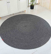 The RAMSHA HOME Black & White Color Round Braided Rug - BR 007 with a spiral pattern lies on a white tiled floor in a bright room, near a cabinet, plant, and large windows with sheer curtains.