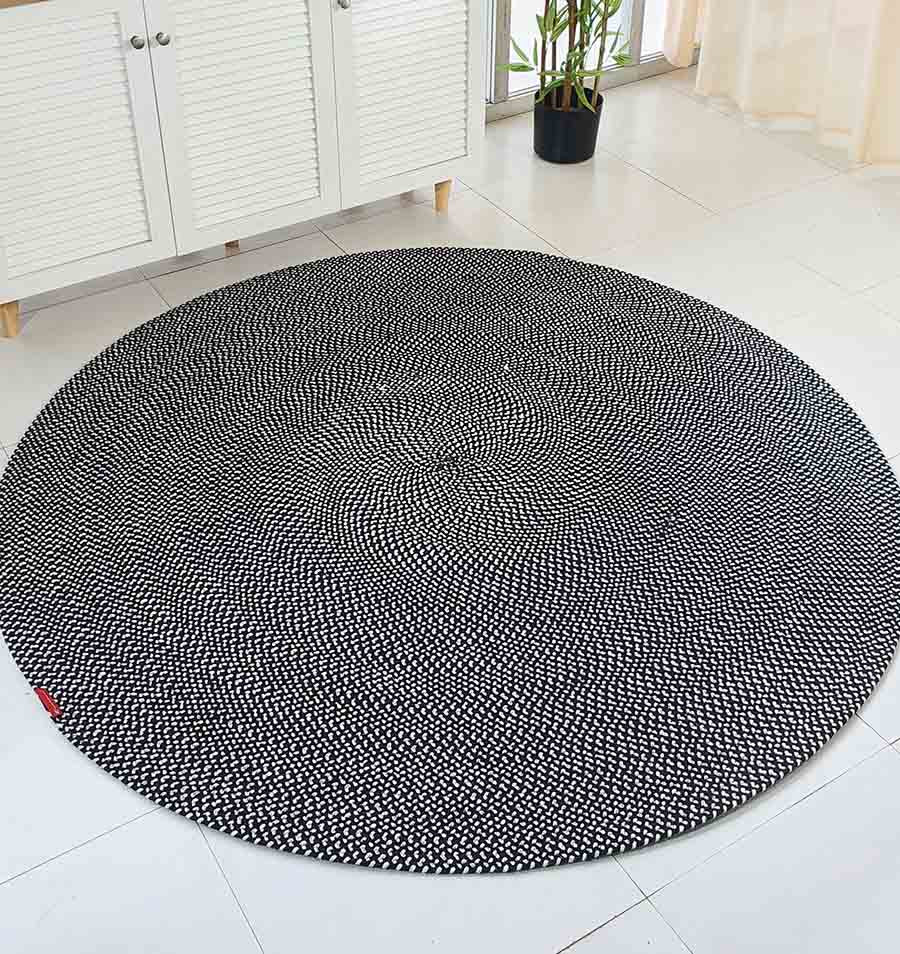 The RAMSHA HOME Black & White Color Round Braided Rug - BR 007 with a spiral pattern lies on a white tiled floor in a bright room, near a cabinet, plant, and large windows with sheer curtains.