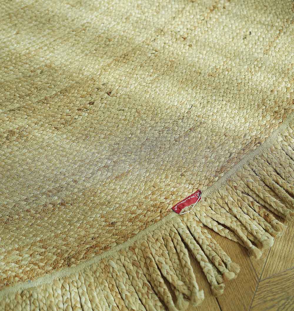 A close-up of the RAMSHA HOME Natural Jute Braided Rug With Fringes BR-015, featuring tasseled fringe and a red fabric tag on a wooden floor. The boho round design in natural fiber brings warmth and style to your space.
