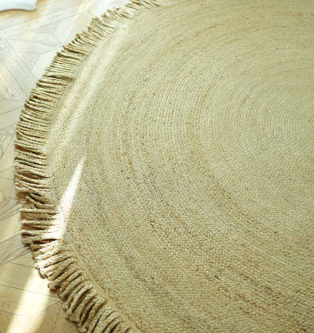 A close-up of the RAMSHA HOME Natural Jute Braided Rug With Fringes BR-015 on a light herringbone wooden floor, sunlight streaming across its textured surface.