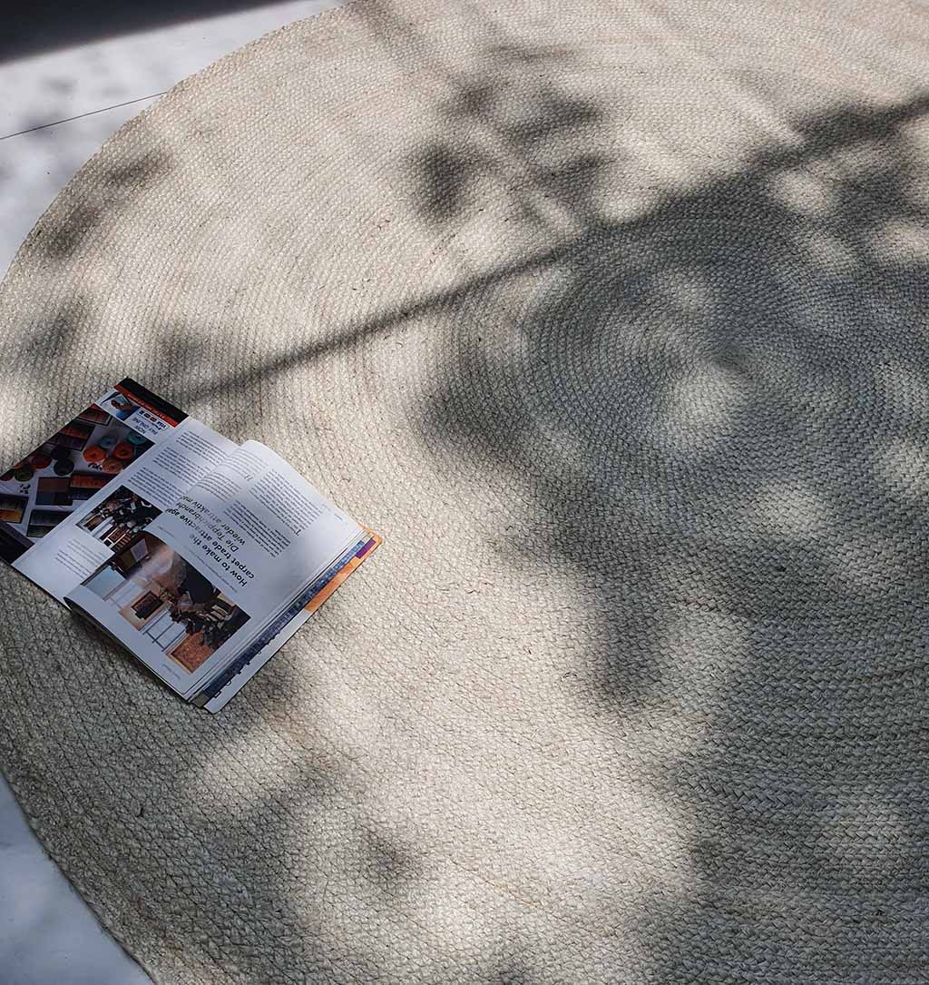 An open magazine rests on the Natural Jute Rug Bleach Color BR-017, with sunlight and leafy shadows playing across its neutral, round surface.