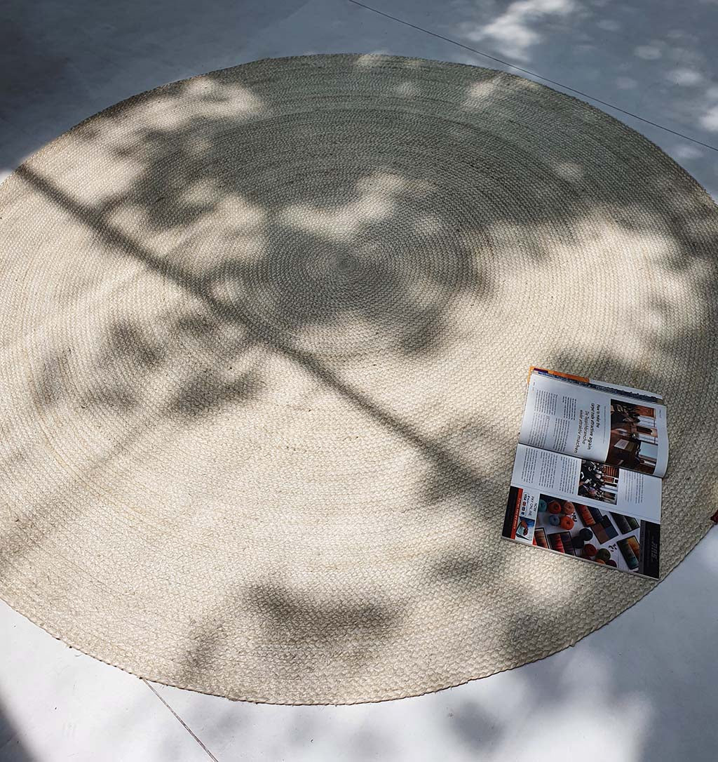 The Natural Jute Rug Bleach Color BR-017 lies on a light floor, its braided texture highlighted by tree-branch shadows. An open magazine rests on the edge, adding cozy style to this naturally chic setting.