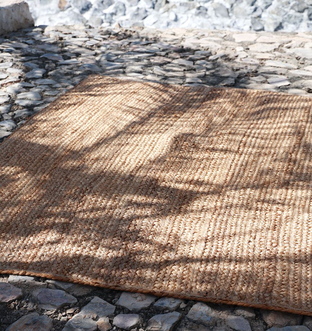 The RAMSHA HOME Stunning Handmade Braided Jute Round Rug - BR 020 (170x240cm) is displayed outdoors on cobblestones, with sunlight filtering through tree branches and casting shadows on its natural fibers.