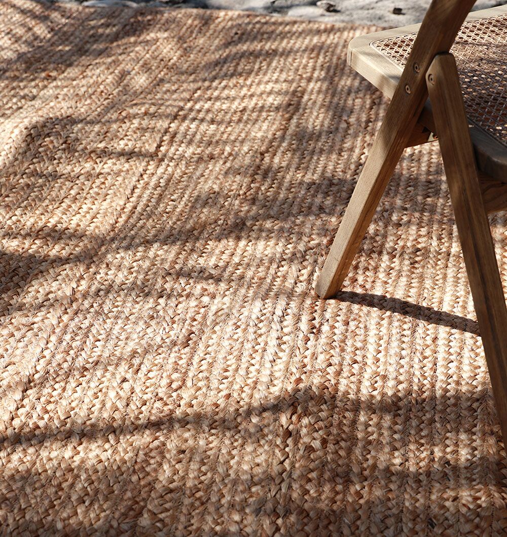 A close-up of the RAMSHA HOME Stunning Handmade Braided Jute Round Rug - BR 020 (170x240cm) with a wooden chair partially visible on the right, bathed in sunlight that creates soft shadows for a warm, cozy feel.