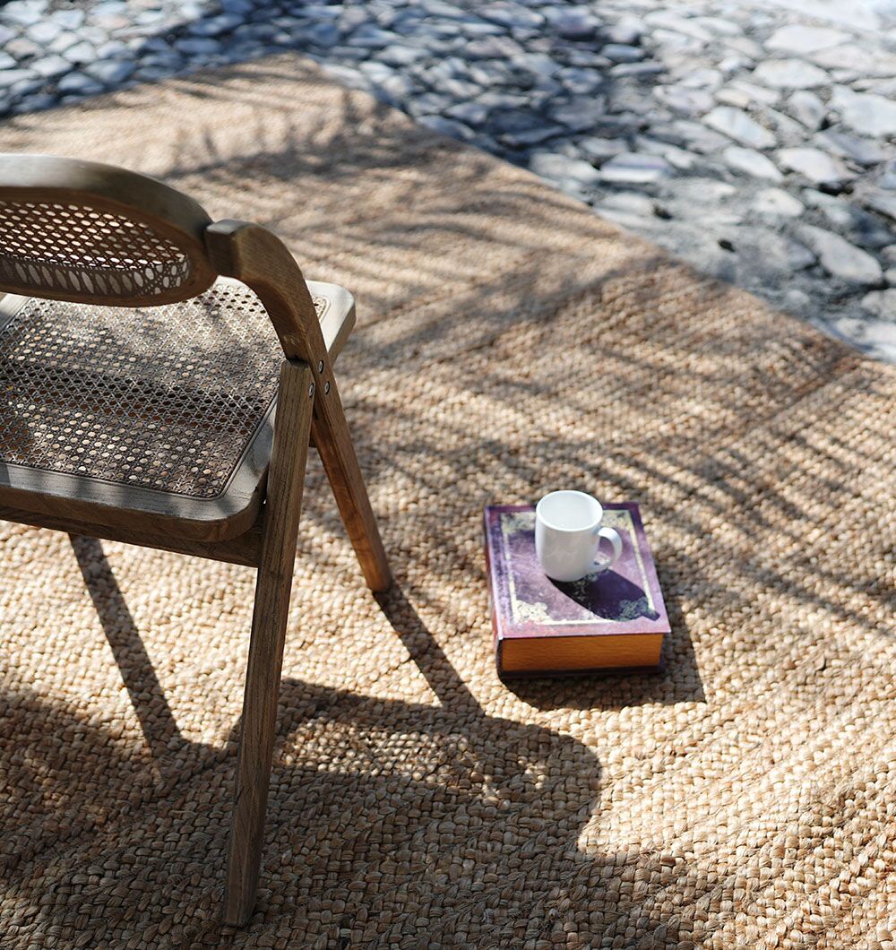 A RAMSHA HOME Stunning Handmade Braided Jute Round Rug (BR 020, 170x240cm) sits outdoors with a wooden chair. Sunlight and palm shadows fall across the tan rug, while a white coffee cup rests on a purple book beside the chair.