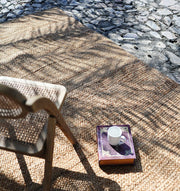 On a sunlit stone patio, a RAMSHA HOME Stunning Handmade Braided Jute Round Rug (BR 020, 170x240cm) sits under a woven chair. A book with a cup rests atop the rug as leaf shadows gently play across the tranquil scene.