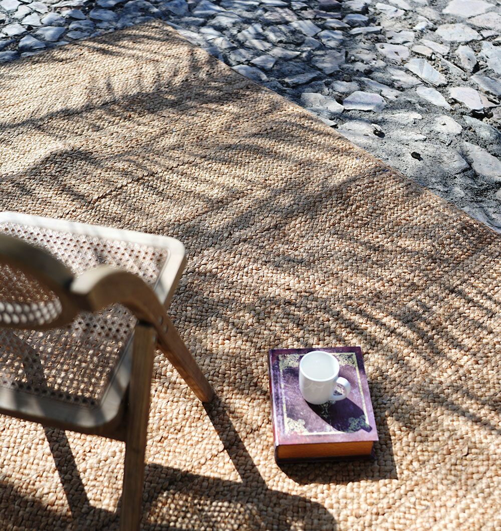 On a sunlit stone patio, a RAMSHA HOME Stunning Handmade Braided Jute Round Rug (BR 020, 170x240cm) sits under a woven chair. A book with a cup rests atop the rug as leaf shadows gently play across the tranquil scene.