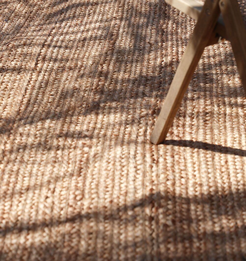 A close-up of the RAMSHA HOME Stunning Handmade Braided Jute Round Rug - BR 020 (170x240cm) shows its rich tan texture in sunlight and shadow; a wooden chair corner appears in the upper right, accentuating this natural fiber rug.