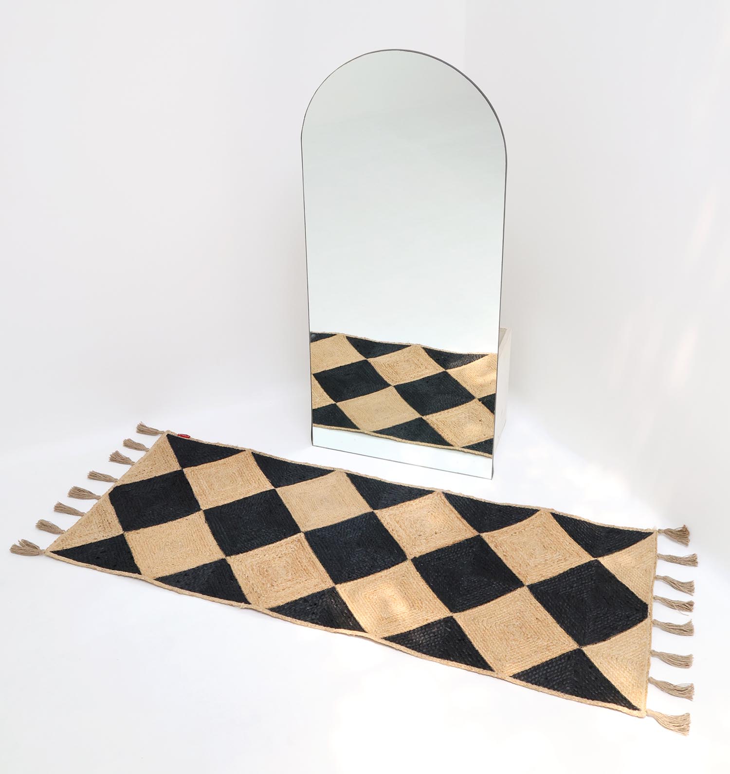A tall, arched mirror stands on a white floor behind the RAMSHA HOME Luxurious Handmade Natural & Black Color Jute Braided Rug with Tassel (BR 030); its black and beige diamond pattern is reflected in the mirror.