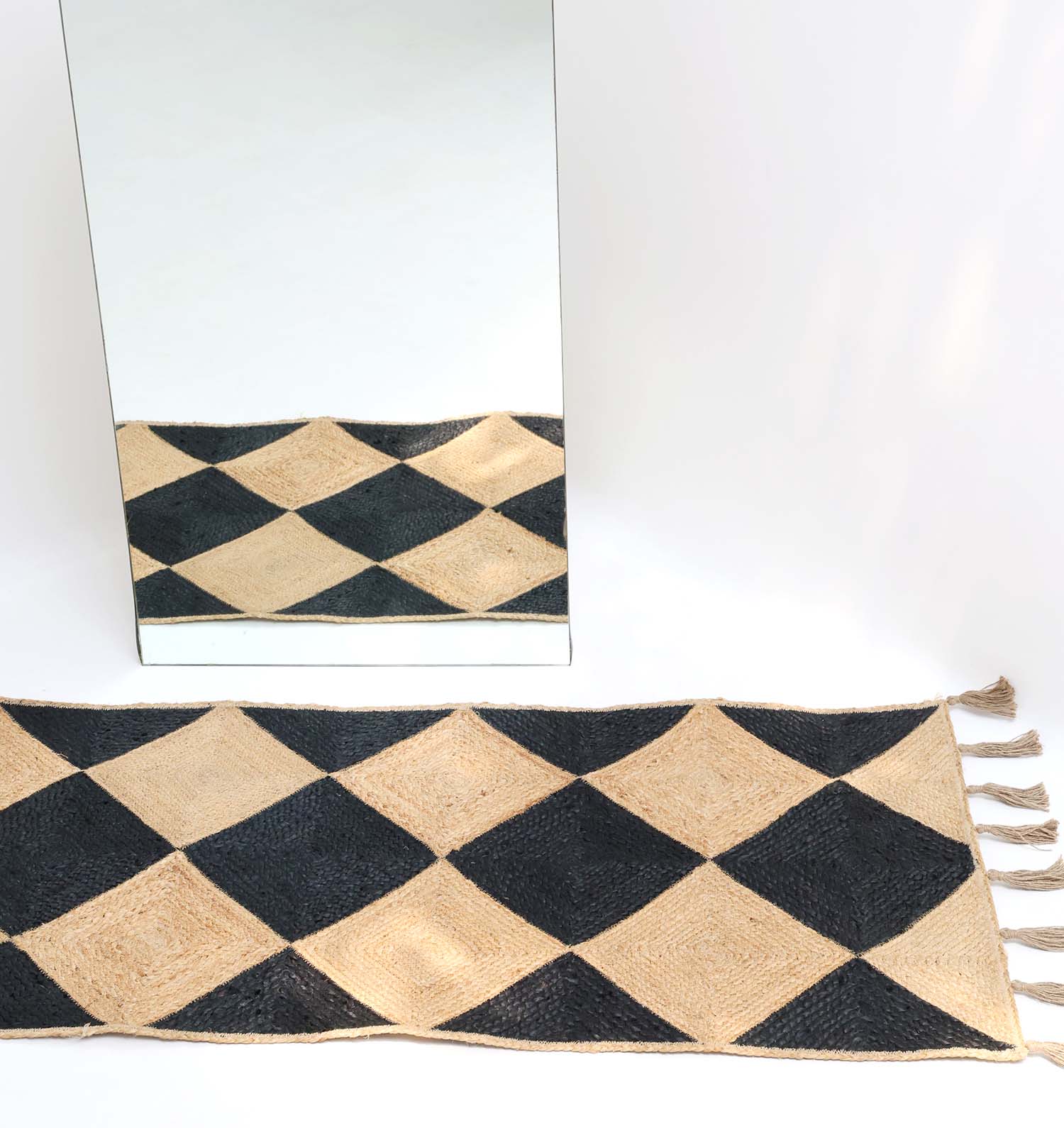 The RAMSHA HOME Luxurious Handmade Natural & Black Jute Braided Rug with Tassel (BR 030) is displayed on the floor, its elegant diamond pattern and tassel detail partially reflected in a large mirror against a white backdrop.