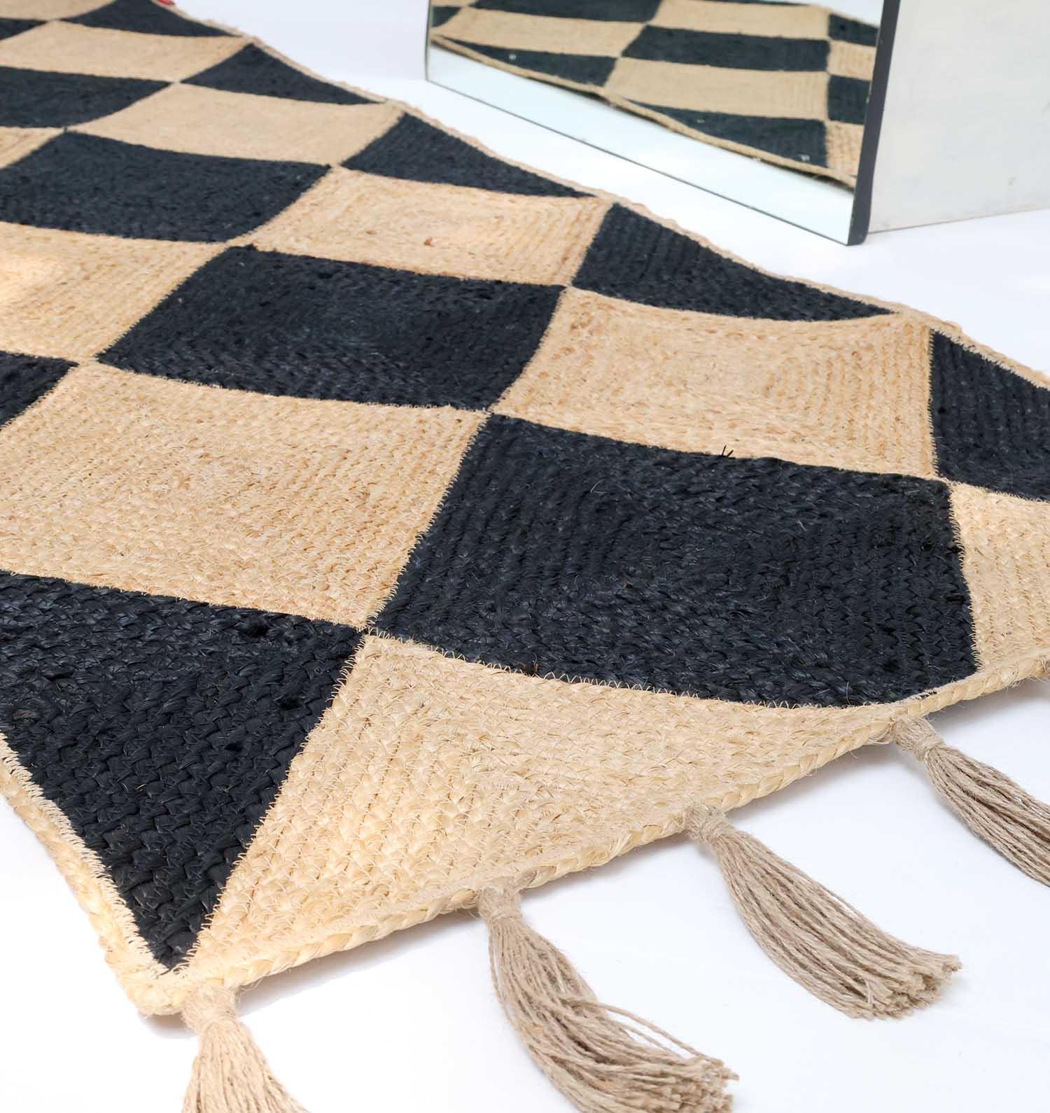 The Luxurious Handmade Natural & Black Color Jute Braided Rug with Tassel- BR 030 by RAMSHA HOME lies on the floor, showcasing its diamond checker pattern and edge tassels. A mirror in the background reflects part of the rug’s design.