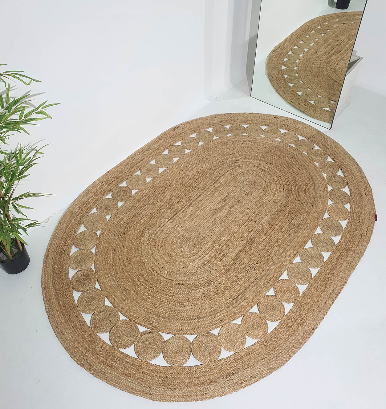 The RAMSHA HOME Beautiful Handmade Jute Floor Braided Rug - BR 032, featuring an oval shape with a border of alternating solid and cut-out patterns, is displayed on a white floor near a green plant and a large mirror reflecting the rug.