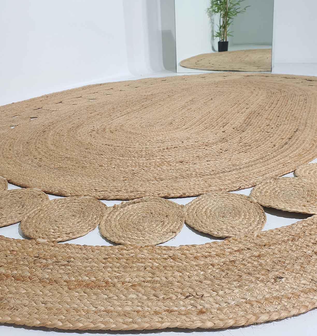 A close-up of the RAMSHA HOME Beautiful Handmade Jute Floor Braided Rug - BR 032 with a circular design and decorative edge is shown on a white floor. A mirror reflects part of the rug and a potted bamboo plant in the background.