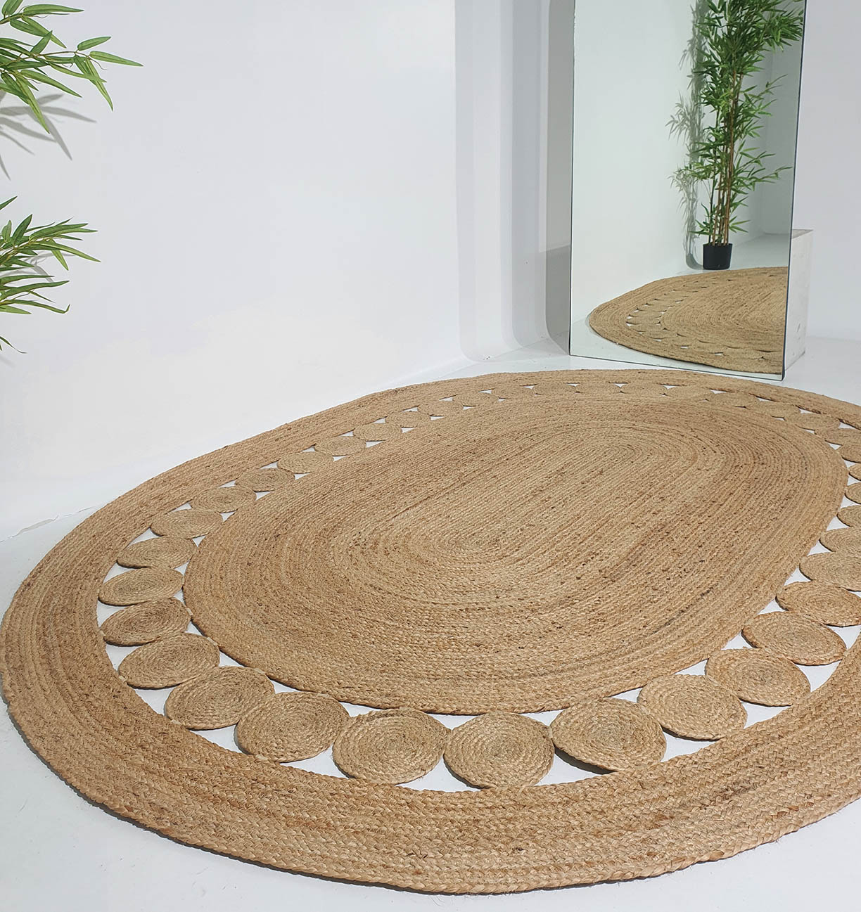 The RAMSHA HOME Beautiful Handmade Jute Floor Braided Rug - BR 032, a large oval piece with a decorative border, rests on a white floor near a tall mirror and green plant, its reflection and the plant partially visible in the mirror.