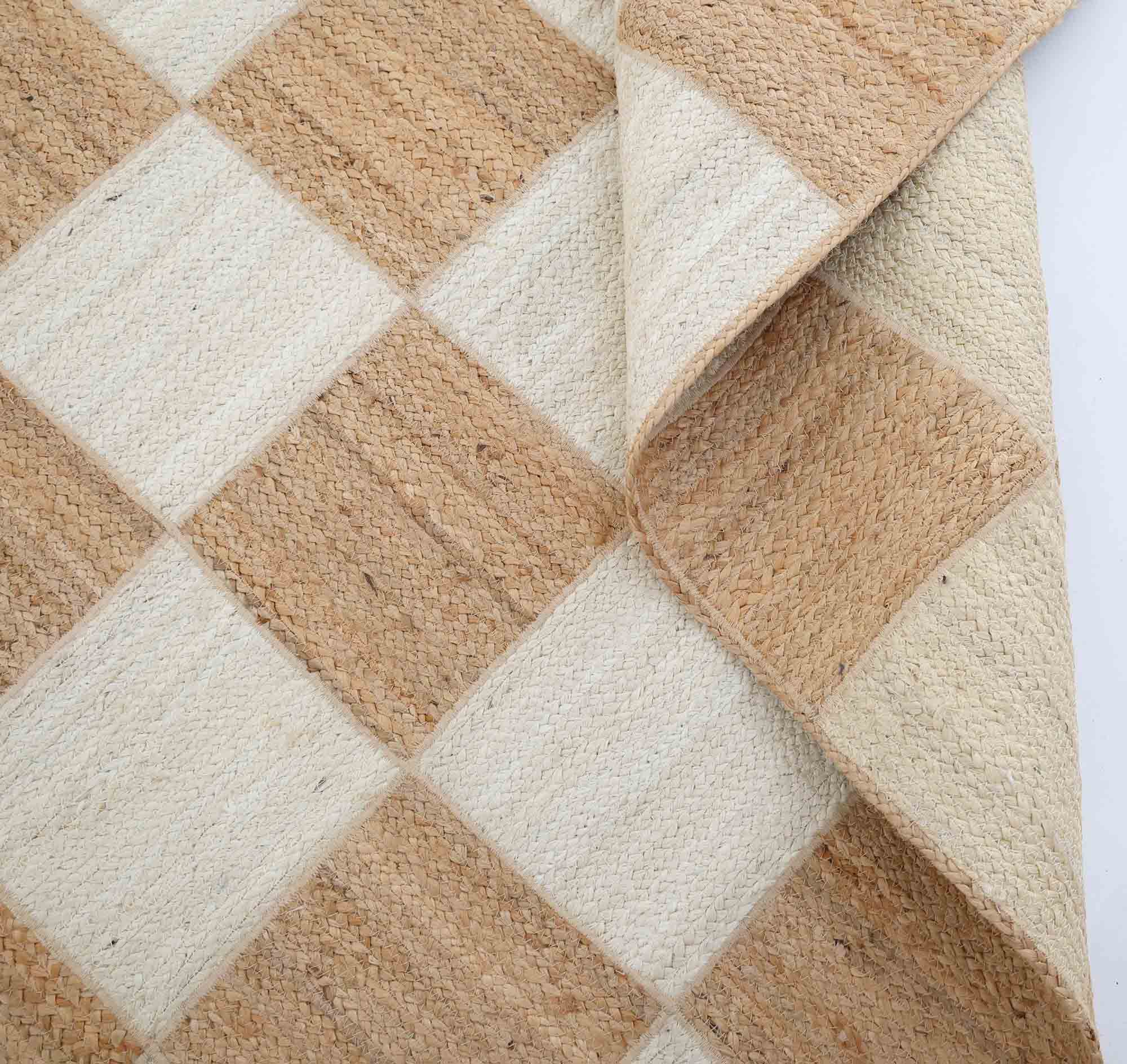 The Stunning Handmade Natural & Bleach Color Outdoor Jute Braided Rug - BR 037 by RAMSHA HOME features a checkered pattern of beige and light brown squares, with one corner folded to display its textured, thick jute craftsmanship.