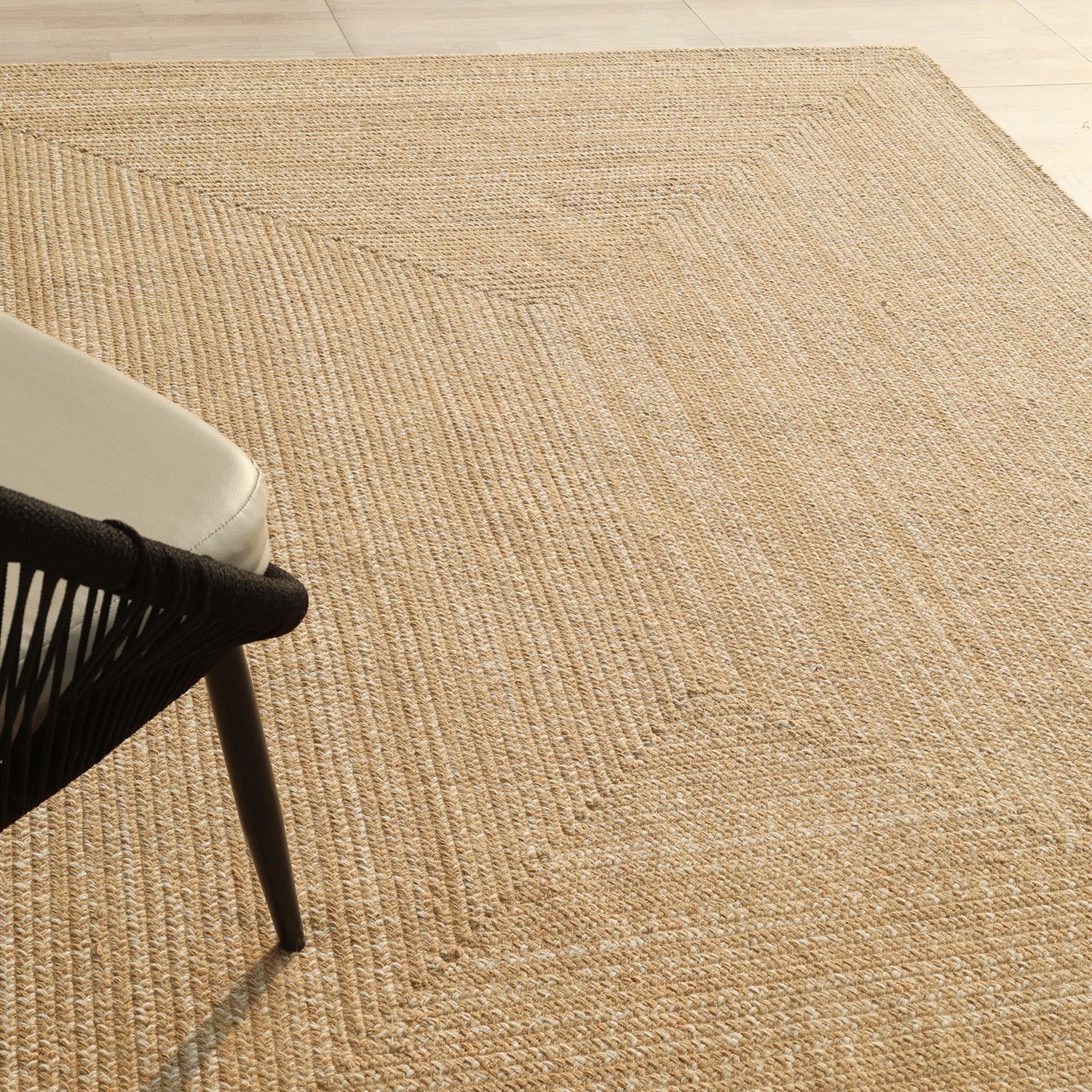 A close-up of the RAMSHA HOME Natural Color Handmade Rectangle Wool Area Braided Rug (BR 052C) on a light floor, with part of a modern chair featuring a black frame and light cushion visible in the corner.