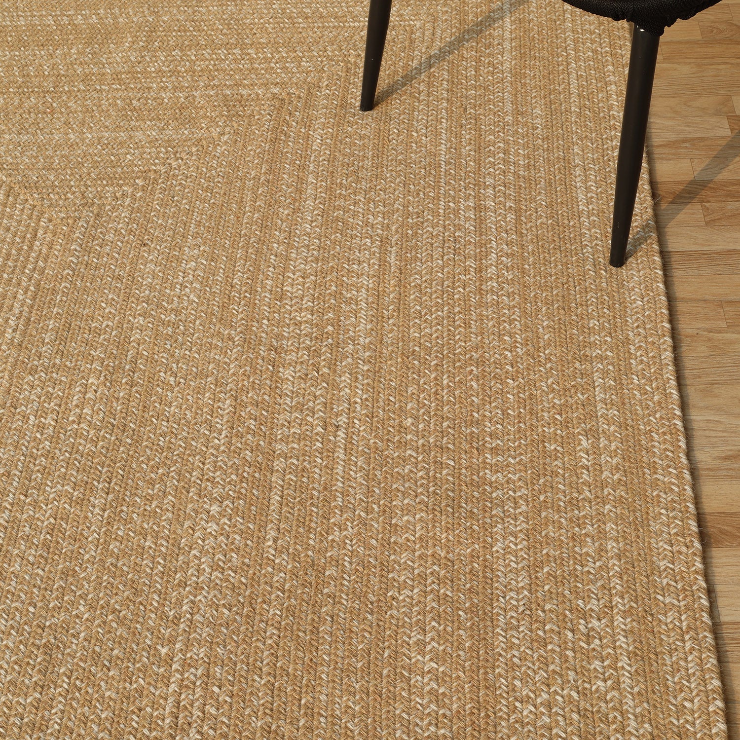 The RAMSHA HOME Natural Color Handmade Rectangle Wool Area Braided Rug - BR 052C lies on a wood floor, with a black chair leg partially visible in the upper right corner.