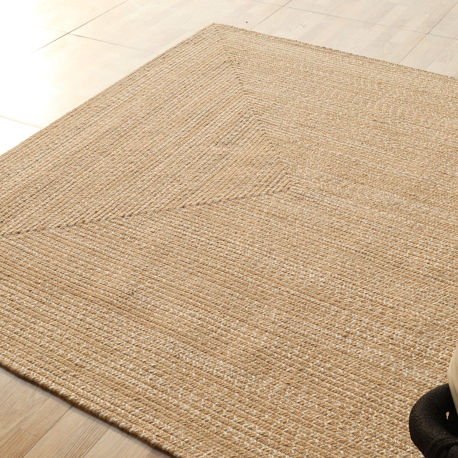A close-up of the RAMSHA HOME Natural Color Handmade Rectangle Wool Area Braided Rug (BR 052C) on a light wood floor, sunlight highlighting its subtle geometric pattern and textured surface for an elegant touch.