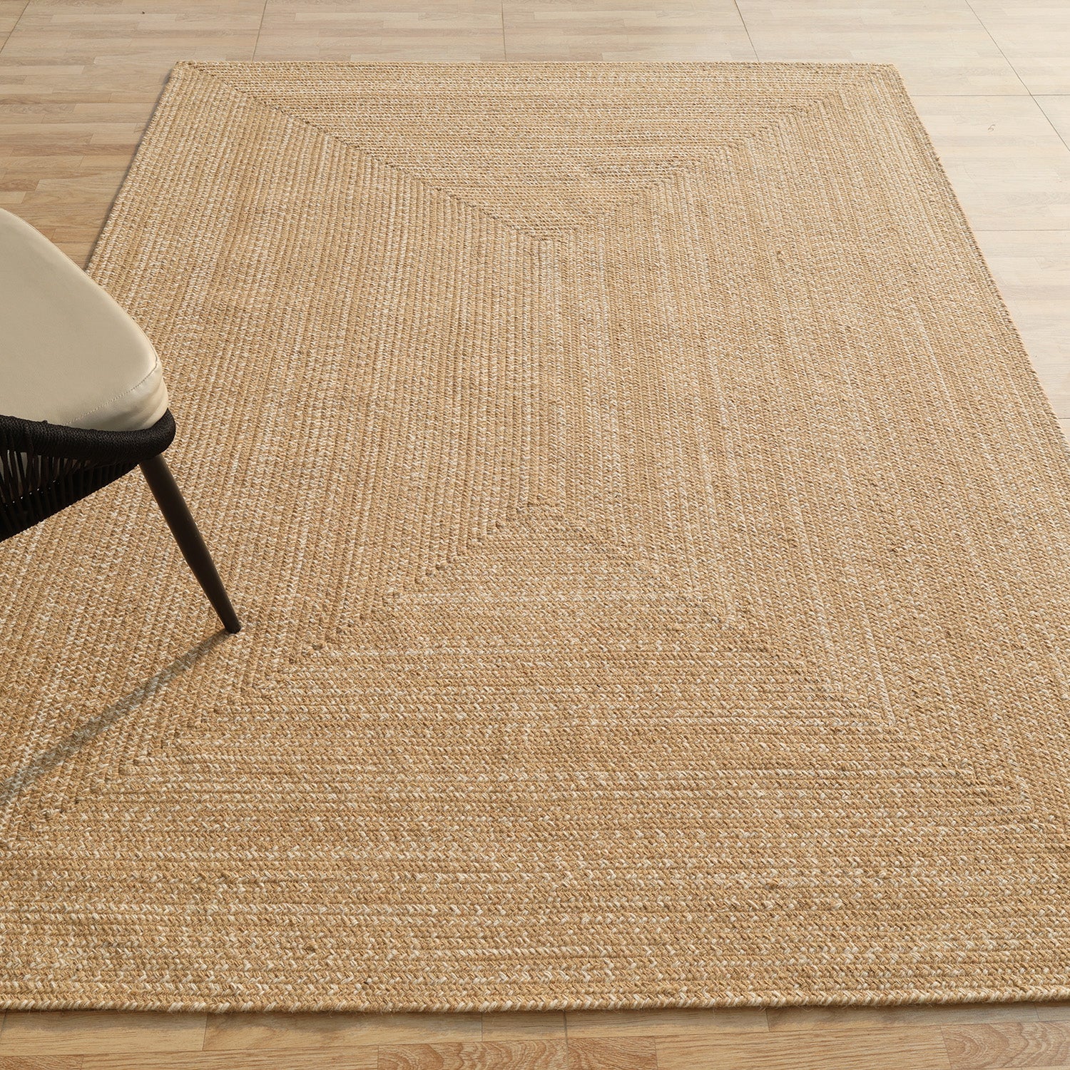The RAMSHA HOME Natural Color Handmade Rectangle Wool Area Braided Rug - BR 052C rests on a light wood floor. In the upper left corner, a black chair with a beige cushion is partly visible.