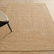 The RAMSHA HOME Natural Color Handmade Rectangle Wool Area Braided Rug - BR 052C rests on a light wood floor. In the upper left corner, a black chair with a beige cushion is partly visible.