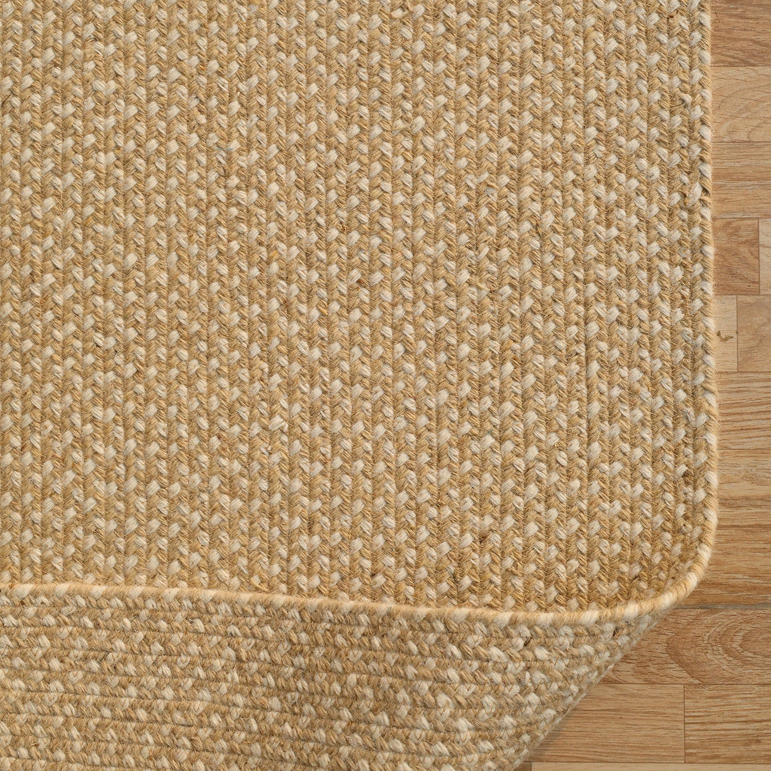 Close-up of the RAMSHA HOME Natural Color Handmade Rectangle Wool Area Braided Rug - BR 053C, shown on a light brown wooden floor. The beige braided rug has rounded edges, a textured pattern, and a slightly lifted corner.