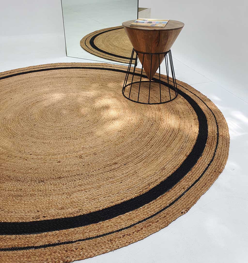 The RAMSHA HOME Handmade Braided Jute Round Rug with Black Border (BR 056) is placed on a white floor beside a small wooden side table with metal legs, with the entire setup reflected in a large wall mirror.