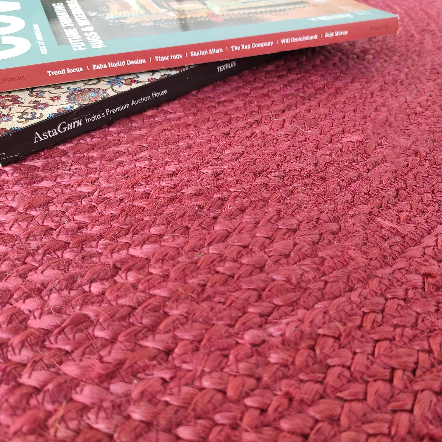 A close-up of the RAMSHA HOME Jute Natural Rug Round Red Color BR-060 (90 cm), with a magazine or catalog titled "AstaGuru: India’s Premium Auction House" resting on it.
