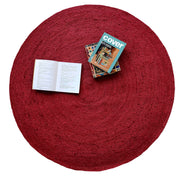 The RAMSHA HOME Jute Natural Rug Round Red Color BR-060 (90 cm) with a braided design is shown from above, featuring a few stacked books and an open book on top, set against a white background.